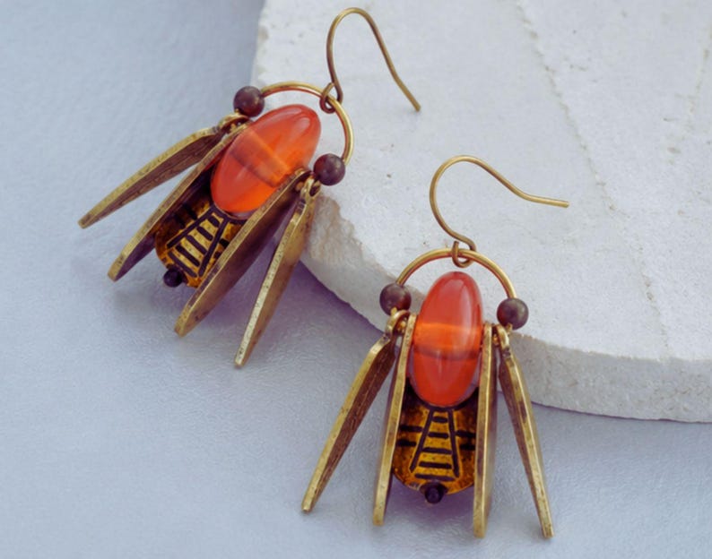 May include: A pair of insect-themed earrings. Each earring features an orange-red oval bead at the centre, framed by brass-toned metal wings and a detailed abdomen. The earrings have a hook closure.