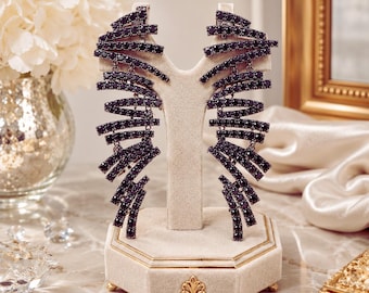 May include: A pair of long, fan-shaped earrings featuring multiple curved, horizontal bars encrusted with small, dark black stones. The earrings are displayed on a beige velvet stand with a gold-toned base and a decorative fleur-de-lis emblem.