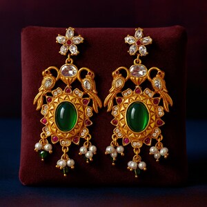 May include: A pair of gold-toned earrings with an oval green gemstone center. The earrings feature bird and floral designs, accented with red and clear gemstones, and dangling pearl clusters. The earrings are displayed on a maroon surface.