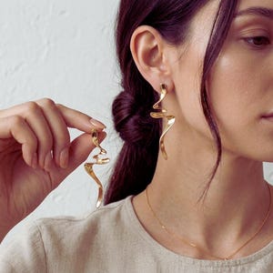 Puede incluir: Pendientes dorados de diseño abstracto. Un pendiente se lleva puesto, el otro lo sostiene una mano. Los pendientes tienen una forma fluida y curvada con un acabado pulido. La joya es una pieza llamativa.