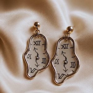 May include: A pair of gold-toned earrings with a surrealist clock face design. Each earring features a distorted, melting clock face with Roman numerals and black hands, suspended from a small gold ball. The background is a cream-colored satin fabric.
