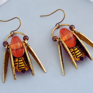 May include: A pair of insect-shaped earrings. Each earring has an orange bead, gold-coloured metal wings, and a detailed black and gold body design. The earrings have a hook closure.