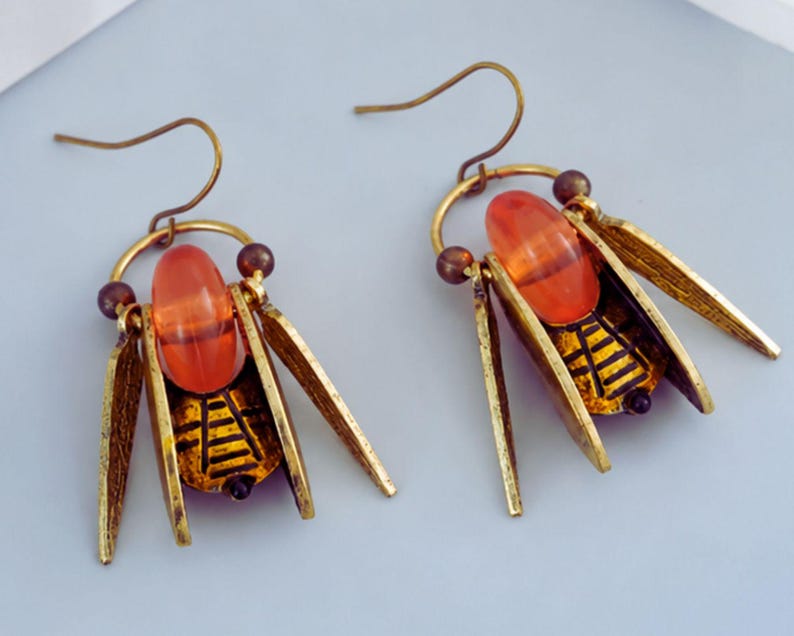 May include: A pair of insect-shaped earrings. Each earring has an orange bead, gold-coloured metal wings, and a detailed black and gold body design. The earrings have a hook closure.