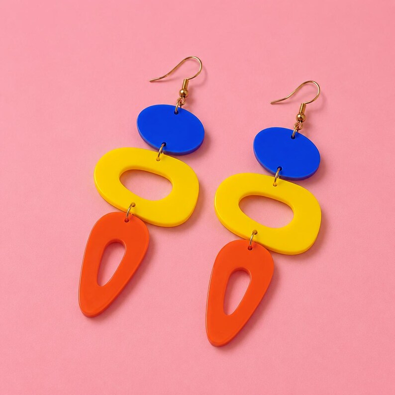 May include: A pair of colorful dangle earrings with a modern design. Each earring features a blue oval, a yellow oval with a hole, and an orange teardrop shape, all connected with gold-colored hooks. The earrings are set against a pink background.