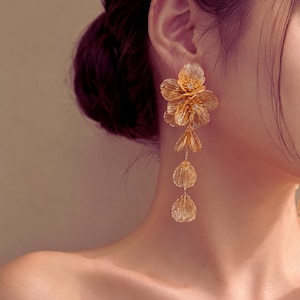 May include: A long gold-toned earring featuring a cluster of textured, petal-like shapes at the top, followed by a vertical drop of three smaller, individual petal-shaped components connected by thin metal links, creating a cascading floral design.