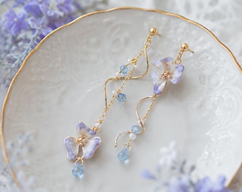 May include: A pair of delicate, gold-toned earrings. Each earring features a light purple and white flower with blue accents and dangling blue teardrop-shaped beads. The earrings are displayed on a white plate with a gold rim, with soft purple flowers in the background.