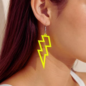 Pode incluir: Brincos em forma de raio amarelo néon. Estes brincos são um acessório divertido e arrojado, com um design simples e delineado. Os brincos penduram-se num gancho de cor prateada.