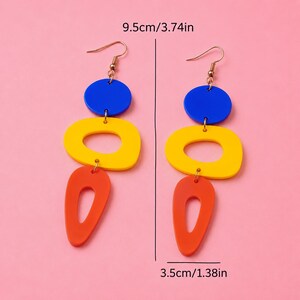 May include: A pair of colorful dangle earrings. Each earring features a blue circle, a yellow oval, and an orange teardrop shape. The earrings are 3.74 inches long and 1.38 inches wide.