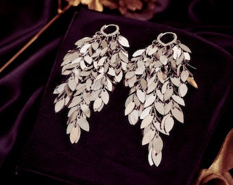May include: Silver leaf-shaped dangle earrings. Each earring features a hoop and multiple tiers of small, flat, leaf-shaped pieces. The earrings are displayed on a dark purple surface, creating a contrast with the bright silver.