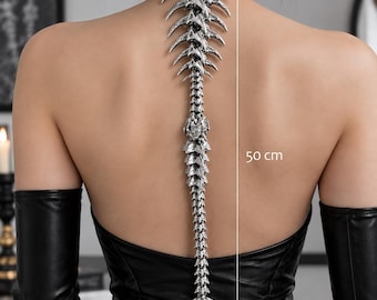 May include: A black leather choker featuring a long, silver-toned spinal column pendant that extends down the back. The metallic vertebrae design is segmented and detailed, measuring 50 cm in total length from the collar to the bottom tip.