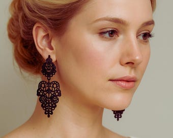 May include: Black filigree earrings with an intricate, openwork design. The earrings are long and dangle, featuring a heart-shaped element. The woman in the image is wearing a white top.