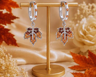 May include: A pair of silver hoop earrings with S925 stamped on the inner band. Each hoop features small orange stones and a dangling maple leaf charm encrusted with orange, yellow, and deep red crystals, suspended from a gold-toned T-bar stand.