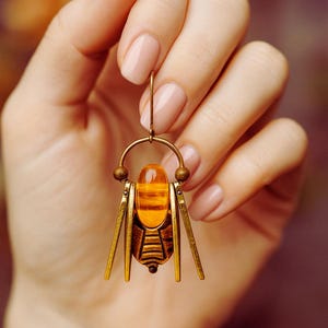 May include: A gold-coloured insect-shaped earring with an amber-coloured central stone. The earring features a detailed design with small decorative elements and is suspended by a gold hook.