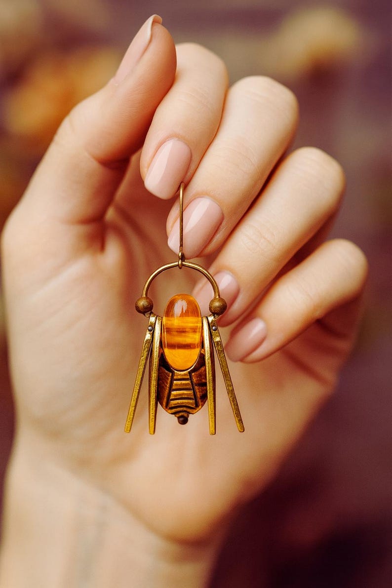 May include: A gold-coloured insect-shaped earring with an amber-coloured central stone. The earring features a detailed design with small decorative elements and is suspended by a gold hook.