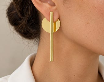 May include: A gold-toned earring featuring a vertical rectangular bar layered over a circular disc. The polished metal surface reflects light, creating a smooth, reflective finish. The design consists of a long, slender bar suspended in front of a flat, round plate, secured through the earlobe.