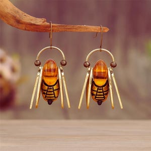May include: Bee-shaped earrings with a golden metal body and amber-coloured oval centres. The wings are detailed. The earrings are suspended from a wooden branch, highlighting their design. The earrings are a unique accessory.
