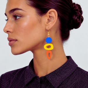 May include: Gold-tone drop earrings with a vibrant, abstract design. The earrings feature a blue circle, a yellow oval, and an orange teardrop shape. The earrings are suspended from a gold hook.