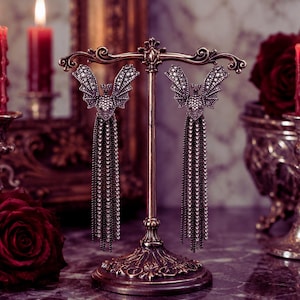 May include: Silver-toned bat earrings with dangling chains of small, sparkling stones. The earrings are displayed on a decorative stand. Red candles and a red rose are in the background. The earrings are a gothic-inspired accessory, suitable for a special occasion.