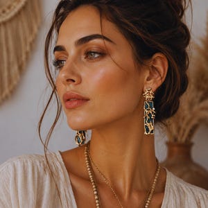 Op de afbeelding: Goudkleurige oorbellen met een rechthoekige, donkergroene inleg en een sierlijk filigraanontwerp. De lange, bungelende oorbellen worden gecombineerd met een gelaagde gouden ketting met een hanger en kralenkettingen. Close-up.