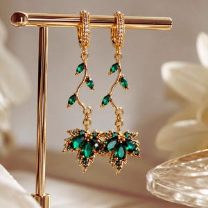 Peut inclure: Une paire de boucles d'oreilles pendantes dorées arborant un motif de vigne délicat avec de petites pierres vertes taille marquise. Les boucles se terminent par un pendentif en forme de feuille d'érable orné de pierres vert foncé et de cristaux.