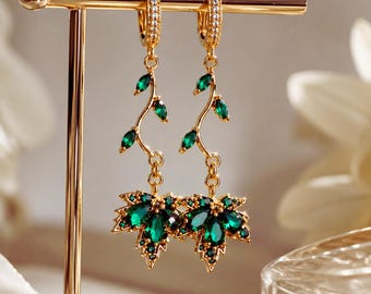 May include: A pair of gold-tone drop earrings featuring a delicate vine design with small marquise-cut green stones. The earrings terminate in a larger maple leaf-shaped pendant encrusted with deep green stones and small clear accent crystals.