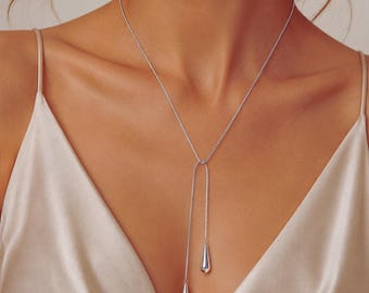 May include: A silver-toned necklace with a delicate chain and two teardrop pendants. The necklace is worn against a cream-colored satin top, creating a contrast in textures and tones. The pendants hang at different lengths.
