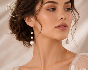 May include: A pair of gold-toned drop earrings featuring a vertical chain design with three white spherical pearls of varying sizes. The earrings hang from the earlobe, with the largest pearl at the bottom and two smaller pearls positioned above it.