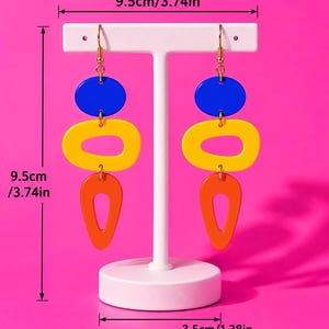 May include: Geometric dangle earrings with blue, yellow, and orange shapes. Each earring is 9.5cm (3.74in) long and 3.5cm (1.38in) wide. The earrings are displayed on a white stand against a bright pink background.