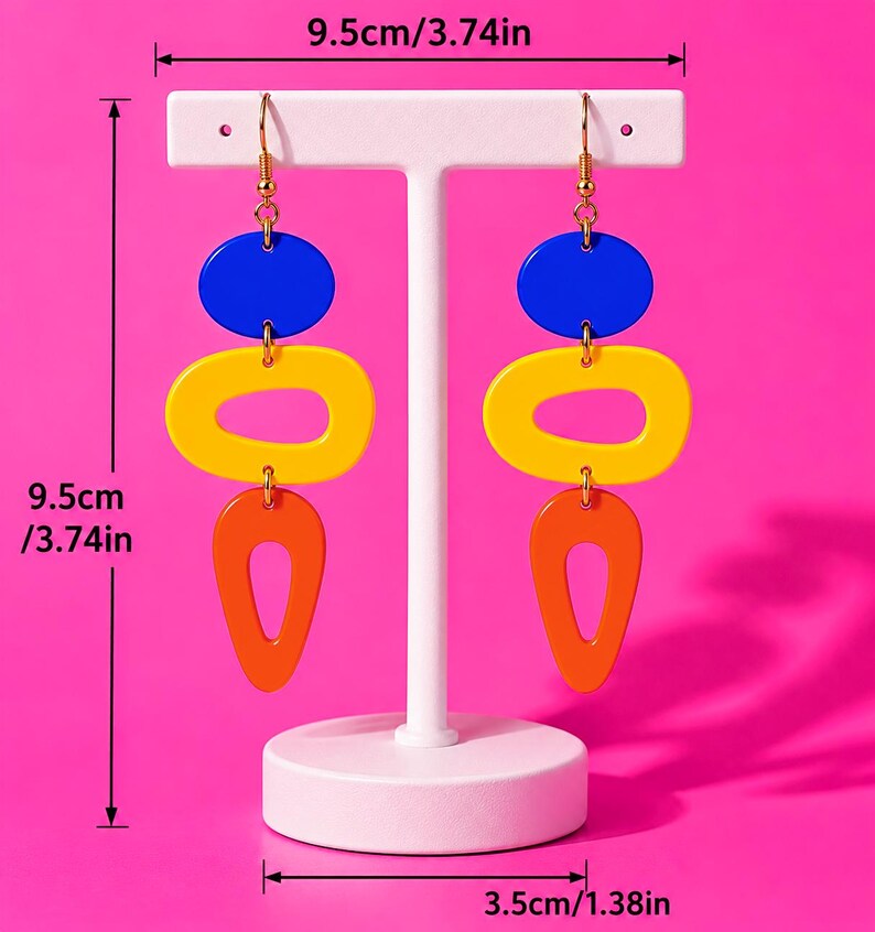May include: Geometric dangle earrings with blue, yellow, and orange shapes. Each earring is 9.5cm (3.74in) long and 3.5cm (1.38in) wide. The earrings are displayed on a white stand against a bright pink background.