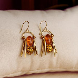 May include: Gold-coloured earrings with a bee motif. Each earring has an amber-coloured oval stone, a detailed bee body, and dangling gold accents. The earrings are presented on a cream-coloured surface.