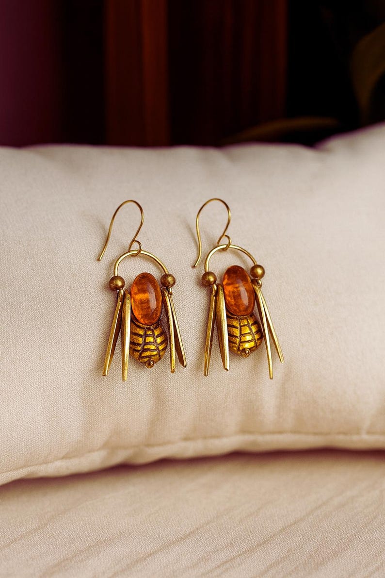May include: Gold-coloured earrings with a bee motif. Each earring has an amber-coloured oval stone, a detailed bee body, and dangling gold accents. The earrings are presented on a cream-coloured surface.