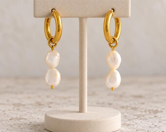 May include: A pair of gold hoop earrings featuring two dangling white baroque pearls on each hoop. The earrings are displayed on a beige T-shaped stand. The hoops have a polished metallic finish and the pearls have an irregular, organic shape.