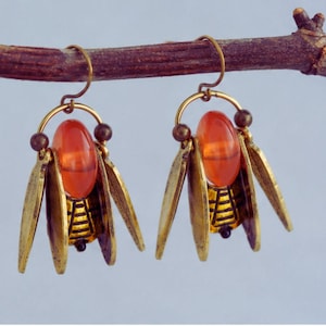 Könnte beinhalten: Ein Paar einzigartige Ohrringe mit einer rötlich-orangen ovalen Perle, die in einem goldfarbenen Metallrahmen hängt. Die Ohrringe haben stilisierte Flügel und kleine braune Perlen, die ein Vintage-Insekten-Design erzeugen.