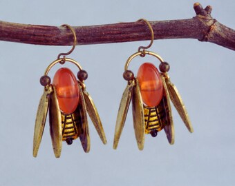 May include: A pair of unique earrings featuring a reddish-orange oval bead suspended within a gold-colored metal frame. The earrings have stylized wings and small brown beads, creating a vintage insect-like design. They hang from a thin, curved gold wire.