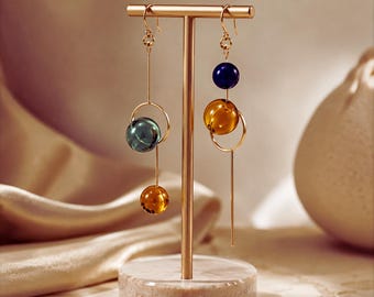 May include: Gold-toned metal earring display stand with two pairs of dangling earrings. Each earring features a combination of colored glass beads and a gold-tone hoop. The beads are in shades of blue, green, and amber.