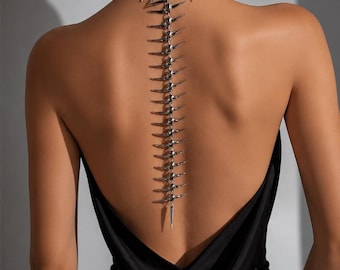 May include: A silver-toned metal body chain necklace featuring a choker collar with sharp, pointed spikes and a long, vertical spine-like extension that drapes down the center of the back, worn over a simple black sleeveless garment.