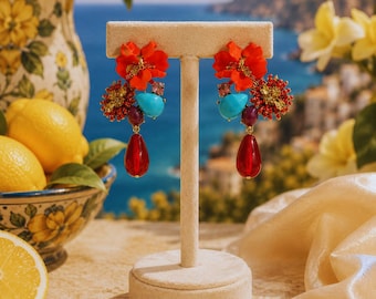 May include: A pair of statement earrings featuring large orange flower petals, clusters of small yellow and red beads, a single turquoise teardrop, a small purple bead, and a large translucent red teardrop pendant hanging from the bottom.