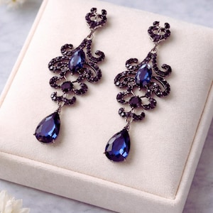 Op de afbeelding: Een paar elegante oorhangers met een donkerblauw en zwart kleurenschema. De oorbellen hebben een druppelvormige blauwe edelsteen aan de onderkant, met ingewikkeld zwart filigraan en kleinere blauwe accenten. De oorbellen worden gepresenteerd op een witte ondergrond.