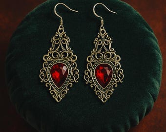 May include: A pair of ornate drop earrings with a vintage aesthetic. Each earring features a large, heart-shaped, deep red gemstone set within an intricate, filigree-style frame. The metal has a warm, antique gold tone, and the earrings are displayed on a dark green velvet surface.