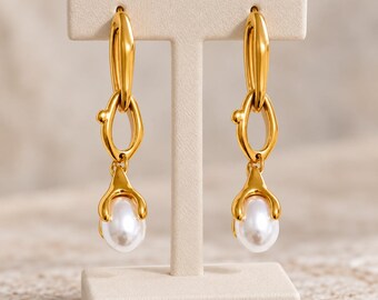 May include: A pair of gold-toned drop earrings featuring an elongated oval link connected to a smaller, organic-shaped link that holds a smooth, white, teardrop-shaped pearl at the base. The earrings are displayed on a T-shaped stand.