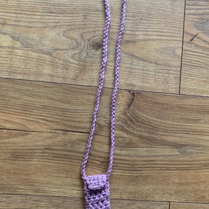 Puede incluir: Una bolsa de ganchillo lavanda con cierre de solapa cuelga de un largo cordón de ganchillo lavanda.