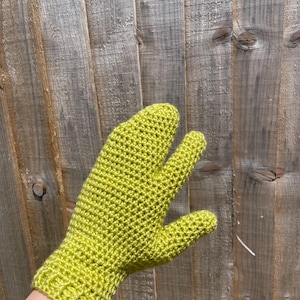 May include: A lime green crochet glove with a unique three-finger design. The glove is made of a textured yarn and features a ribbed cuff. The glove is shown against a weathered wooden background.