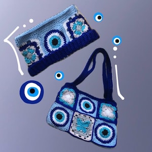 May include: A crocheted blue headband and handbag set. The headband has a dark blue band and is decorated with light blue and white floral patterns and a central eye design. The handbag has a matching design with a butterfly motif.