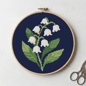 May include: A cross-stitch embroidery of lily of the valley flowers in a wooden hoop. The design features white bell-shaped flowers, green leaves, and a dark blue background. The hoop is approximately 7 inches in diameter.