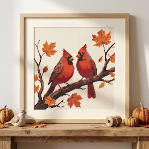 May include: A framed cross-stitch embroidery featuring two red cardinal birds perched on a brown tree branch with orange and yellow maple leaves. The artwork is displayed in a light-colored wooden frame, showcasing a detailed pixelated design.