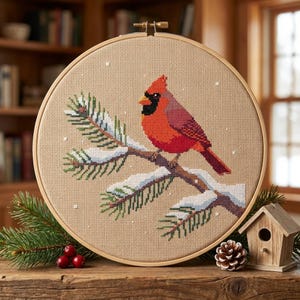 May include: A circular wooden embroidery hoop containing a cross-stitch design of a bright red cardinal bird perched on a snow-covered pine branch against a tan fabric background, with small white dots representing falling snow across the scene.