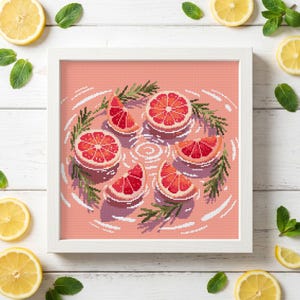Puede incluir: Un bordado en punto de cruz enmarcado con un diseño de arte de píxeles de cinco rodajas de pomelo y ramitas de romero verde flotando en el agua. El fondo es de un color rosa salmón sólido y el marco es un borde cuadrado blanco.