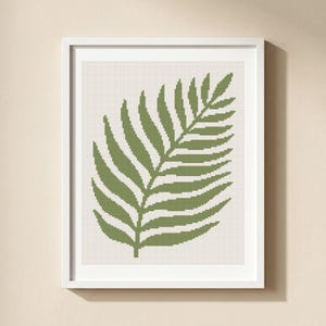 May include: A framed print featuring a green fern leaf design. The leaf is rendered in a pixelated style against a cream-colored background. The artwork is encased in a white frame, suitable for wall decor.