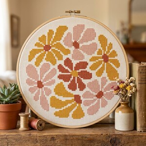 May include: A circular wooden embroidery hoop containing a cross-stitch pattern of stylized flowers in shades of mustard yellow, dusty pink, and terracotta orange on a white fabric background, featuring a repeating floral motif across the surface.