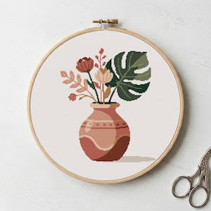 Könnte beinhalten: Kreuzstich-Stickerei einer Vase mit Blumen und einem großen Blatt. Die Vase ist rotbraun mit einem dekorativen Muster. Die Blumen sind in Rot-, Rosa- und Cremetönen gehalten, und das Blatt ist grün. Die Stickerei befindet sich in einem Holzreifen.